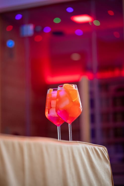 Two vibrant cocktails with ice on white tablecloth, blurred neon-lit bar background at Suite Apartment Taormina