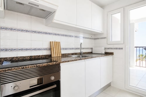 Cocina moderna blanca con encimera de granito, electrodomésticos de acero inoxidable y vistas al mar en el apartamento Riurau Home Calpe