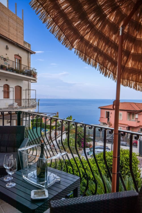 Taormina suite balcony with sea view, wine glasses, and Mediterranean coastline beyond