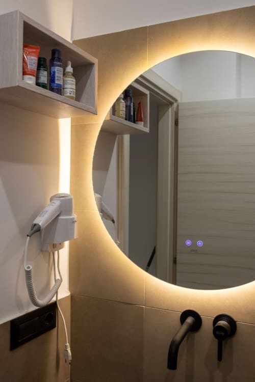 Modern bathroom in Taormina suite with illuminated round mirror, wall-mounted sink, black faucet, and storage shelf with