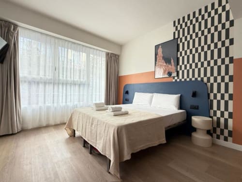Modern suite at Groma Rooms hostel in Zaragoza with blue headboard, checkered wall art, and large windows