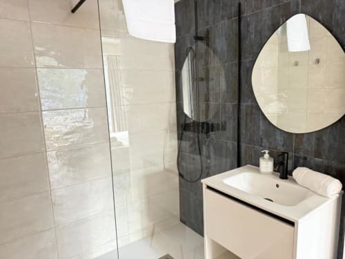 Modern bathroom at Groma Rooms hostel in Zaragoza with white sink, round mirror, dark tile shower area, and glass partition
