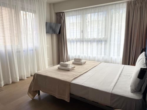 Bright suite at Groma Rooms hostel in Zaragoza with white bedding, rolled towels, floor-to-ceiling windows, and neutral
