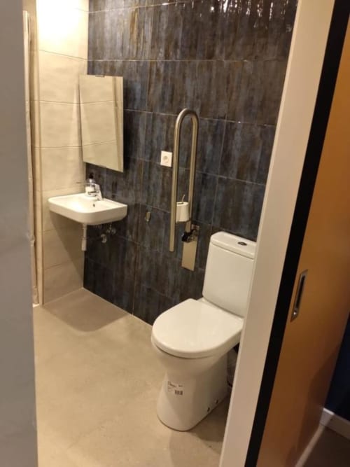 Accessible bathroom at Groma Rooms hostel in Zaragoza with toilet, sink, grab bars, and dark tile shower area