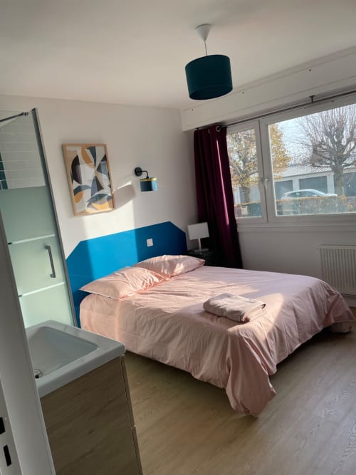 Modern bedroom at Mattbnb Metz with blue headboard, pink bedding, pendant light, and city view windows