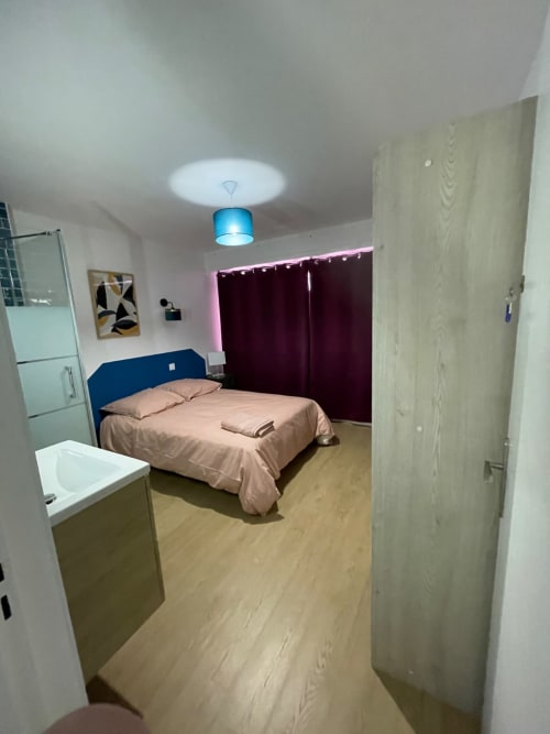 Chambre moderne chez Mattbnb à Metz avec tête de lit bleue, linge de lit beige, rideaux bordeaux et salle de douche attenante