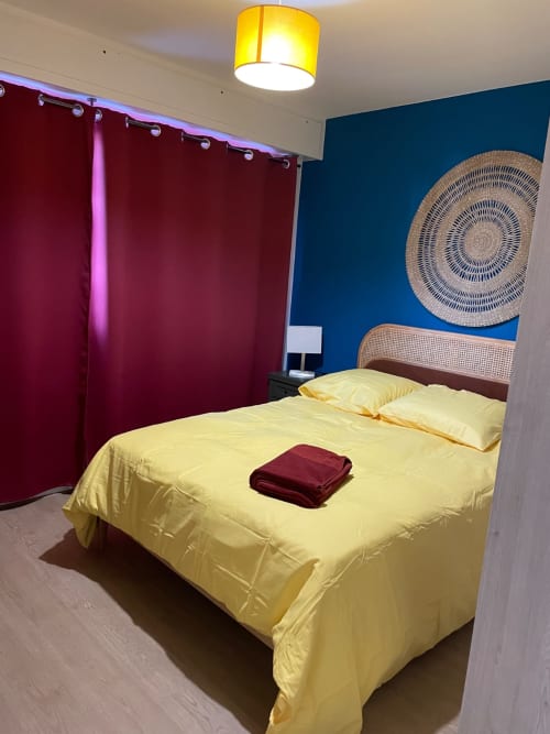 Chambre lumineuse chez Mattbnb Metz avec literie jaune, rideau bordeaux, mur d'accent bleu et art mural circulaire tressé