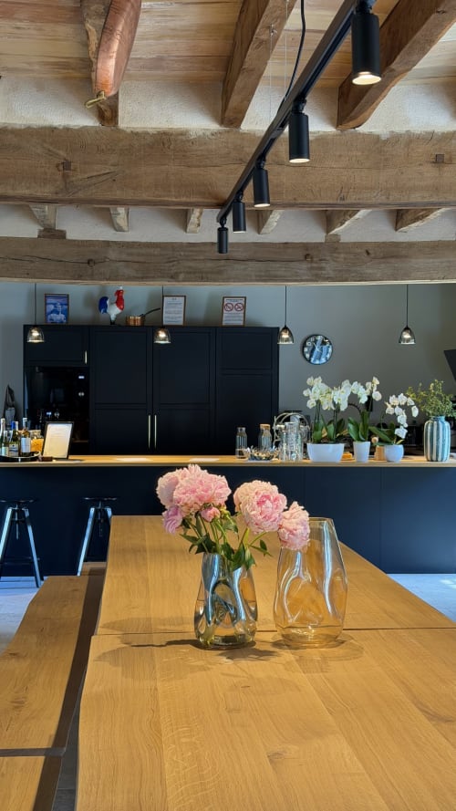 Espace repas du Cloffy BnB avec poutres en bois, cuisine noire, pivoines roses en vases de verre et luminaires suspendus modernes