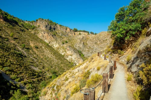 Barandilla de madera a lo largo de sendero montañoso con cañón rocoso y vegetación verde cerca de Hospedería Puerta a los Cahorros en Monachil