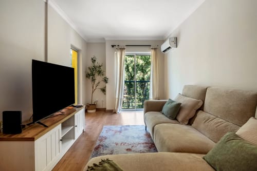 Sala luminosa em Atlantic Places Albufeira com sofá bege, TV e vista para o jardim da varanda