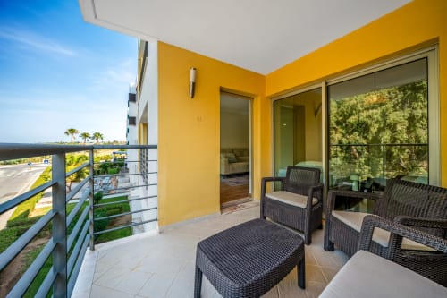 Varanda ensolarada em Atlantic Places Albufeira com paredes amarelas, móveis de vime preto e vista para o jardim