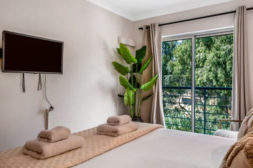 Quarto moderno no Atlantic Places Albufeira com vista para o jardim, cama em bege, TV na parede e planta em vaso