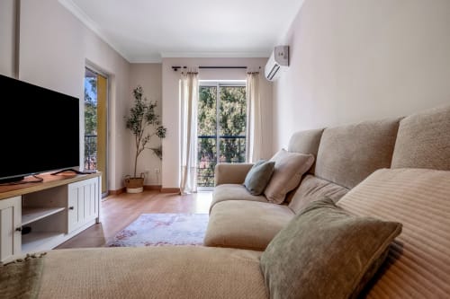 Sala de estar luminosa no Atlantic Places Albufeira com sofá bege, TV e vistas do jardim pelas portas da varanda