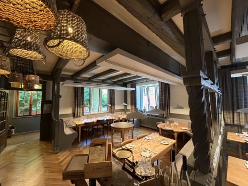 Élégante salle à manger de l'Hôtel l'Ange à Niedermorschwihr avec poutres en bois, lampes en osier et tables dressées près de grandes fenêtres
