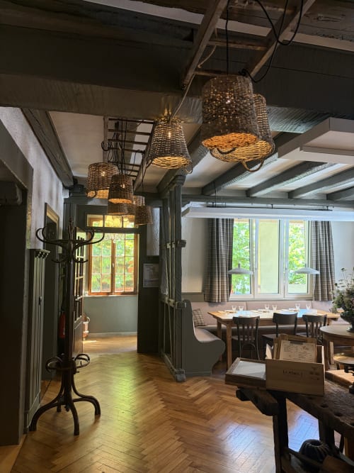 Salle à manger rustique de l'Hôtel l'Ange avec luminaires tissés, tables en bois et grandes fenêtres donnant sur les jardins