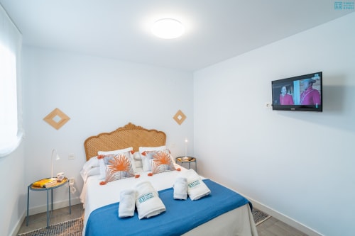 Dormitorio moderno con cama azul, cabecero de mimbre, almohadas naranjas, TV montada en pared y paredes blancas en Alhaurín Loft City Center
