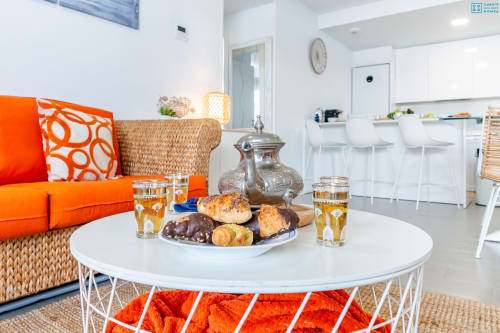 Sala de estar moderna en apartamento de lujo en Alhaurín con sofá naranja, mesa redonda blanca, pastelería y cocina abierta