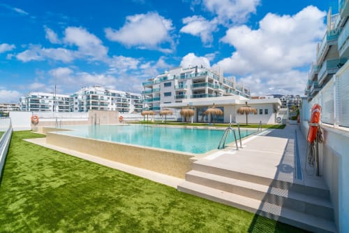 Piscina de lujo en Alhaurín Loft con agua turquesa, playa de arena, edificios modernos blancos y cielo azul