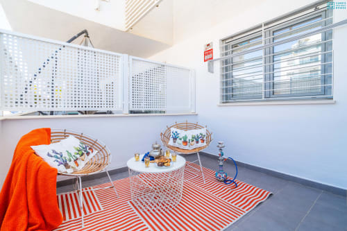 Moderno espacio de vida con alfombra de rayas rojas y blancas, sillas tejidas con plantas en maceta y ventanas luminosas en Alhaurín