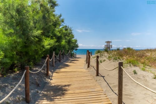 Pasarela de madera con barandillas de cuerda que conduce a una playa de arena con mar turquesa, torre de salvavidas y vegetación verde