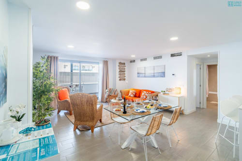 Sala de estar de apartamento moderno y luminoso con acentos naranjas, mesa de comedor, sillas de mimbre, balcón con vista al mar y diseño contemporáneo