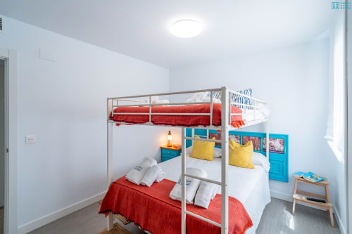 Dormitorio infantil luminoso con cama alta, ropa de cama roja, marco blanco, almohadas amarillas y unidad de almacenamiento azul en Alhaurín