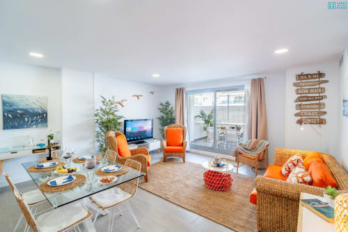 Sala moderna de apartamento frente al mar con muebles naranjas, mesa de comedor, TV, balcón con vista al mar y decoración costera