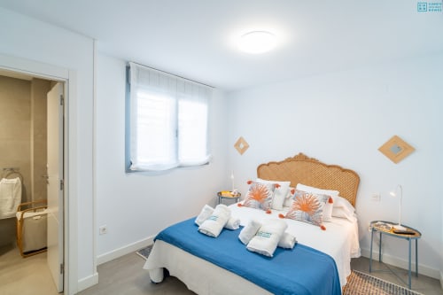 Dormitorio luminoso con cabecero de ratán, manta azul, cojines naranjas y vistas al mar en Alhaurín Loft