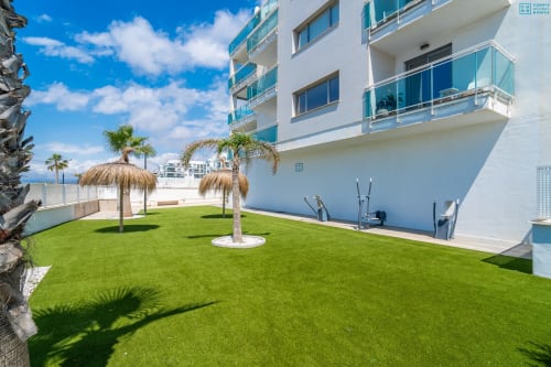 Patio de apartamento de lujo frente al mar con césped cuidado, palmeras y edificio moderno blanco en Alhaurín Loft City