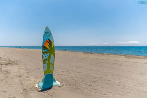 Tabla de surf colorida clavada en la arena de la playa de Alhaurín con mar turquesa y cielo despejado