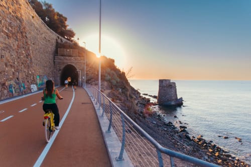 Ciclista sulla passeggiata costiera al tramonto a Marina di Andora, Italia, con vista mare e architettura storica in pietra