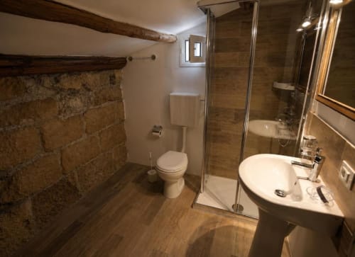 Bagno moderno con parete in pietra, doccia in vetro, lavabo bianco su piedistallo e pavimento in legno presso Politeama Central Suite