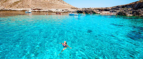 Snorkelista nuota nelle acque turchesi cristalline della spiaggia Marausa con barche e scogliere rocciose