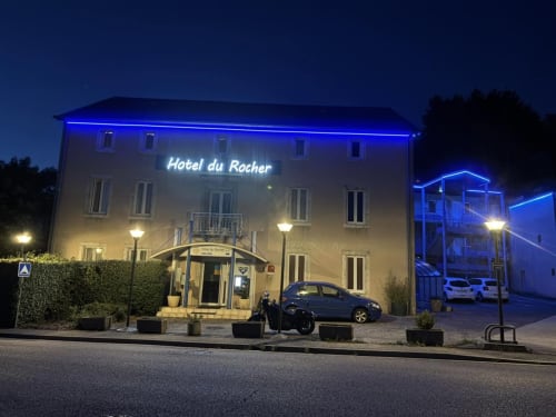 Hôtel Du Rocher la nuit, illuminé en bleu néon avec son entrée à Le Caylar, France