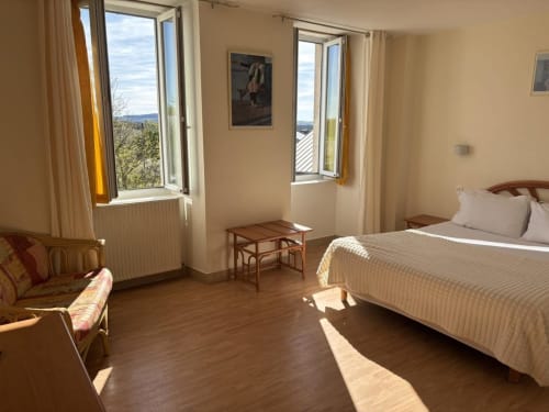 Chambre lumineuse à l'Hôtel Du Rocher avec lit, coin salon et vues pittoresques sur la campagne depuis les fenêtres