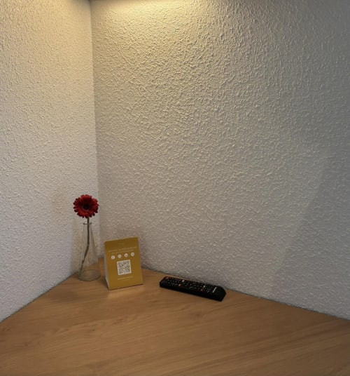 Zimmerecke im Hôtel Du Rocher mit strukturierten weißen Wänden, Holzboden, rote Gerbera in einer Vase, goldene Box und Fernbedienung