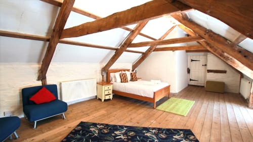 Spacious attic bedroom with exposed wooden beams, white walls, blue seating, wooden bed, hardwood floors, and skylight at