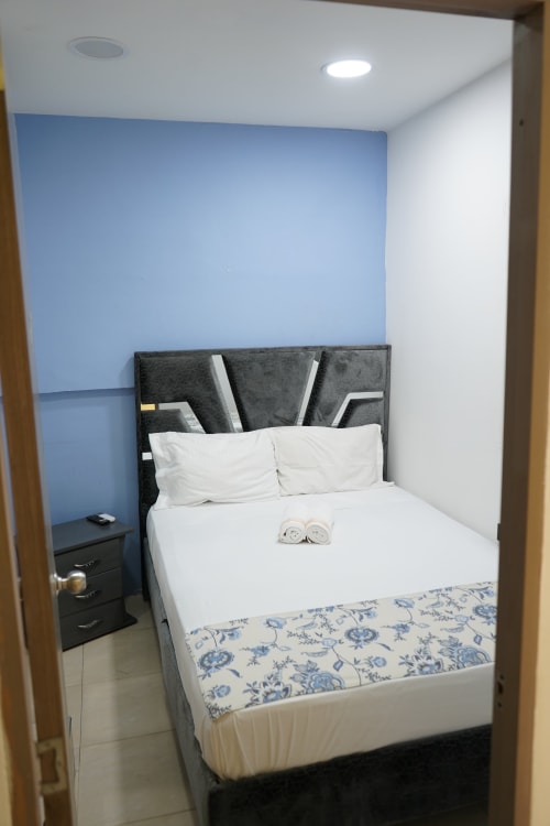 Habitación con cama doble en hotel Casa Mango de Cali con pared azul, cabecero gris, sábanas blancas y toallas enrolladas