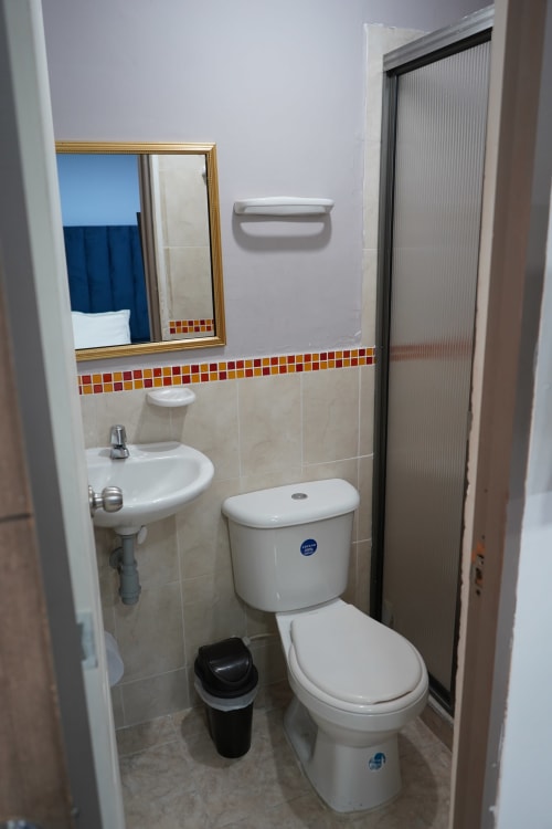 Baño compacto del hotel Casa Mango Cali con inodoro blanco, lavamanos pedestal, espejo marco dorado y azulejos coloridos