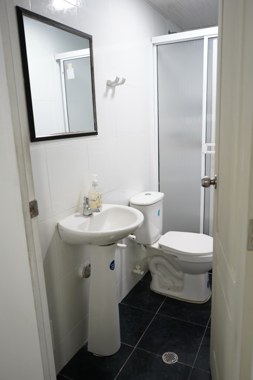 Baño moderno y limpio del hotel Casa Mango en Cali con fixtures blancos, piso de azulejos negros y espejo enmarcado