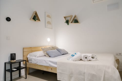 Dormitorio minimalista en Stay Gest Valencia con cama de madera, ropa de cama blanca, toallas, repisas flotantes con plantas y detalles en negro