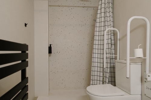 Baño accesible en Stay Gest Valencia con barras de seguridad blancas, inodoro, cortina de ducha de rejilla, toallero eléctrico negro