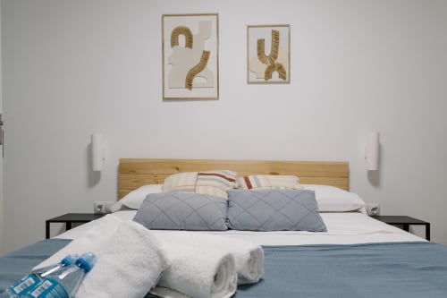 Habitación moderna en Stay Gest Valencia con cabecero de madera, ropa de cama blanca, almohadas grises, toallas enrolladas y pared minimalista