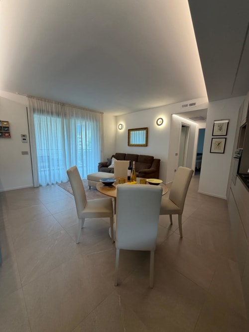 Sala da pranzo moderna nell'appartamento La Carezza di Arco con tavolo rotondo, sedie bianche, soggiorno con divano marrone e giardino
