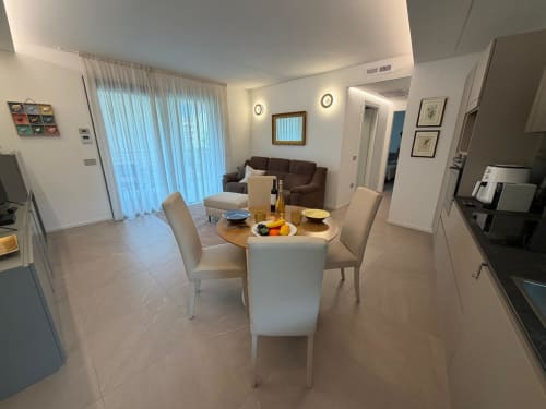 Ampio soggiorno con tavolo rotondo, sedie color crema, divano marrone, cucina e vista giardino