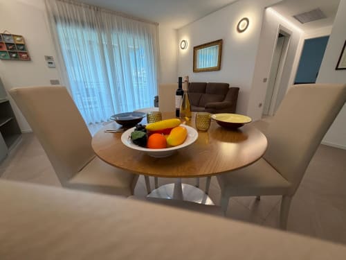 Moderna zona pranzo nell'appartamento La Carezza di Arco con tavolo rotondo in legno, sedie bianche, centrotavola di frutta fresca e giardino