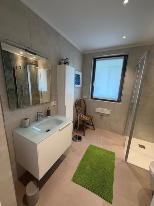 Bagno moderno dell'appartamento La Carezza di Arco con lavabo bianco, doccia walk-in e finestra con vista sul giardino