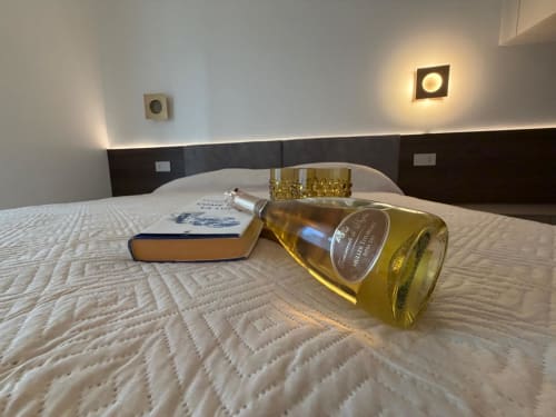 Camera a La Carezza di Arco con bottiglia di vino, libro e bicchiere sul letto, testata scura, illuminazione incassata