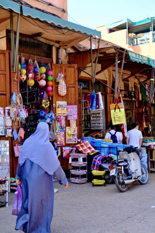 Vibrant Marrakech medina market stall with colorful hanging balls and textiles, woman in blue djellaba shopping near parked