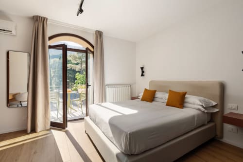 Camera luminosa con vista montagna, letto beige, cuscini arancioni, balcone ad arco e dettagli in legno